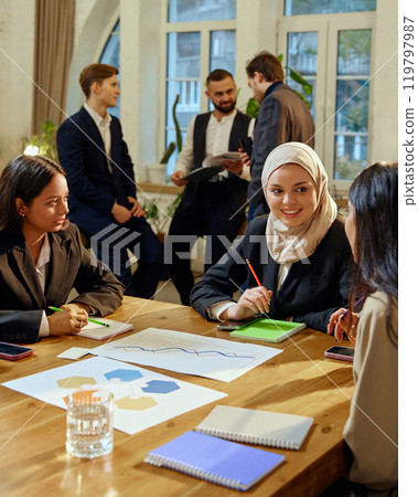 Multicultural female colleagues engaged in discussion during corporate strategy meeting. Other team, male members talking in the background. Multicultural female colleagues engaged in discussion during corporate strategy meeting. Other team, male members talking in the background. 119797987