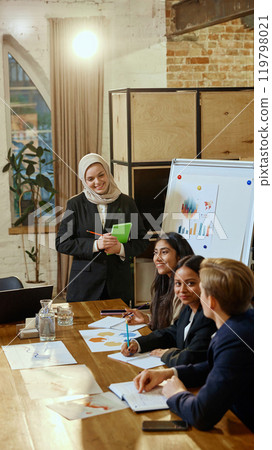 Woman in hijab presenting financial data and trends to diverse group of colleagues. 119798021