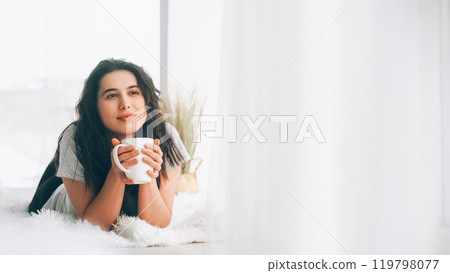 Morning coffee. Enjoying rest. Lazy sunday. Relaxed happy woman with cup of tea lying floor near window in modern light cozy apartment copy space. Morning coffee. Enjoying rest. Lazy sunday. Relaxed happy woman with cup of tea lying floor near window in modern light cozy apartment copy space. 119798077