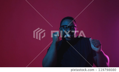 Winner dance. Victory celebration. Overjoyed funky excited smiling man satisfied with success in neon light isolated on dark red background empty space. 119798080