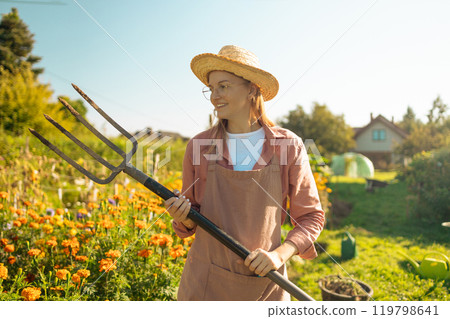 Beautiful Caucasian woman farmer walking in field early in morning with pitchfork. Farming lifestyle. Female holding fork and going to work outdoor. Girl in village, working on the field.  119798641