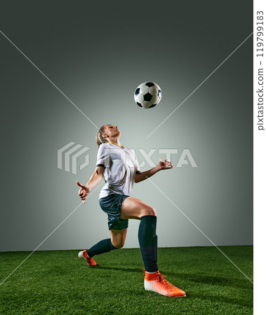 Full-length image of focused and determined young woman, soccer player in motion on field, training, hitting ball with chest. Grey background. 119799183