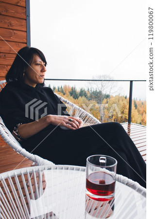 Young woman sitting on terrace with wine in cups on foggy forest background. Tranquil relax in mountains. Thougftful girl on autumn mountains background. Vacations in Ukraine. Chill in Carpathians. Young woman sitting on terrace with wine in cups on foggy forest background. Tranquil relax in mountains. Thougftful girl on autumn mountains background. Vacations in Ukraine. Chill in Carpathians. 119800067