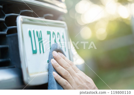 A car owner carefully wipes down their license plate, reflecting Thai beliefs in lucky numbers on vehicle plates and the attention given to maintaining auspicious symbols 119800110