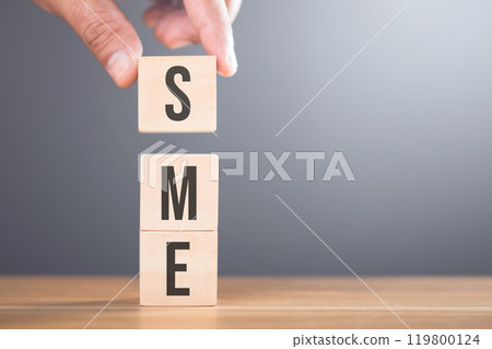 A hand arranging wooden blocks with the letters 'SME' on a table, set against a gray background. Concept of small and medium-sized enterprises, highlighting their importance in the business landscape A hand arranging wooden blocks with the letters 'SME' on a table, set against a gray background. Concept of small and medium-sized enterprises, highlighting their importance in the business landscape 119800124
