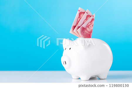 A hand holding a 100 Yuan note, dropping it into a white piggy bank, with Chinese Yuan currency placed on a table against a light blue. Saving and managing money in China's financial context A hand holding a 100 Yuan note, dropping it into a white piggy bank, with Chinese Yuan currency placed on a table against a light blue. Saving and managing money in China's financial context 119800130