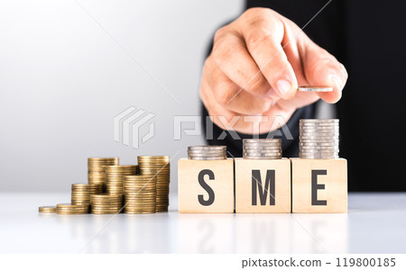 Hand stacking coins with 'SME' written on a wooden block and more coins on the table. Represents financial growth of small and medium-sized enterprises, emphasizing investment and money management Hand stacking coins with 'SME' written on a wooden block and more coins on the table. Represents financial growth of small and medium-sized enterprises, emphasizing investment and money management 119800185