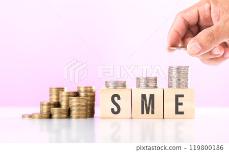 Hand stacking coins with 'SME' written on a wooden block and more coins on the table. Represents financial growth of small and medium-sized enterprises, emphasizing investment and money management 119800186
