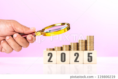 Stack of coins, a wooden block labeled '2025,' and a hand holding a magnifying glass. Examining financial plans and investments for the upcoming year, highlighting careful analysis and planning 119800219