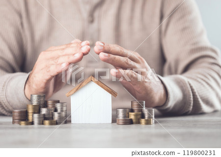 Businessman making a protective hand gesture over a stack of coins and a house model. Image represents the concept of asset protection, emphasizing safeguarding financial investments and property 119800231