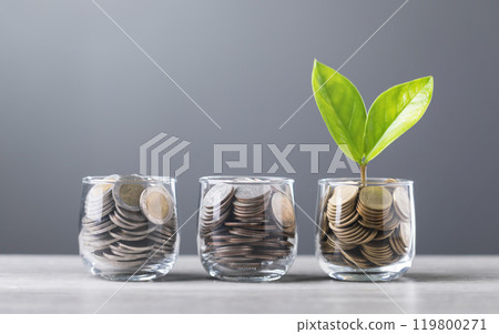 Glass jar filled with coins, with a small seedling growing in the center, placed on a table against a dark grey backdrop. The image represents the concept of saving and financial growth for future 119800271