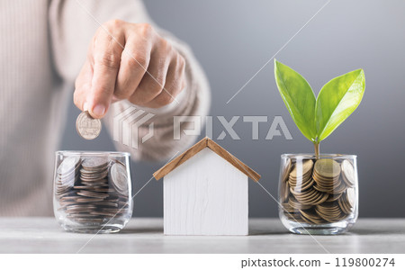 Man drops coins into a glass jar, with stacks of coins and a wooden house model beside him. Emphasizes the concept of saving money for future house investments and financial stability Man drops coins into a glass jar, with stacks of coins and a wooden house model beside him. Emphasizes the concept of saving money for future house investments and financial stability 119800274
