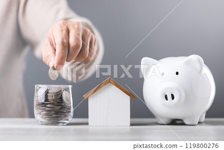 Man drops coins into a glass jar, with piggy bank and a wooden house model beside him. Emphasizes the concept of saving money for future house investments and financial stability 119800276