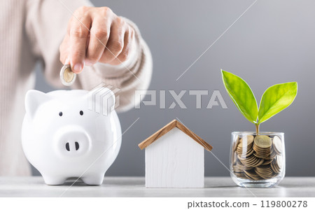 A Man drops coins into white piggy bank, glass jar of coin and a wooden house model beside him. Emphasizes the concept of saving money for future house investments and financial stability A Man drops coins into white piggy bank, glass jar of coin and a wooden house model beside him. Emphasizes the concept of saving money for future house investments and financial stability 119800278