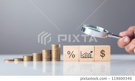 Wooden block labeled interest icons sits beside a stack of coins, while a hand holds a magnifying glass over them. The image symbolizes the scrutiny of taxes and financial analysis 119800348