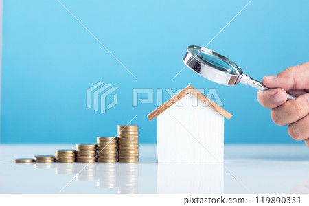 A wooden house model next to a stack of coins, with a hand holding a magnifying glass over them. The image represents the concept of analyzing and calculating home maintenance and repair costs 119800351