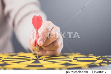 A businessman's hand precisely placing a red dart in the bullseye of a yellow dartboard. The image symbolizes focus, precision, and achieving goals with strategic accuracy. 119800370