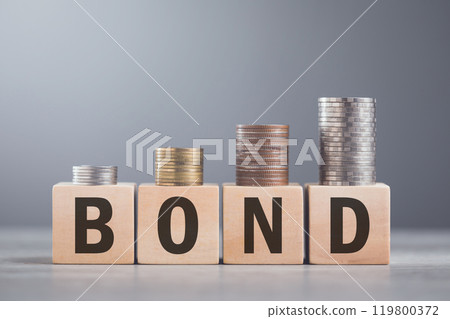 A hand stacking coins on a wooden block labeled 'BOND,' The image reflects the steady and structured growth associated with bond investments, emphasizing stability and financial planning 119800372