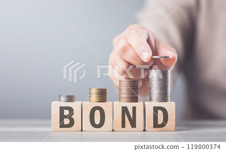 A hand stacking coins on a wooden block labeled 'BOND,' The image reflects the steady and structured growth associated with bond investments, emphasizing stability and financial planning A hand stacking coins on a wooden block labeled 'BOND,' The image reflects the steady and structured growth associated with bond investments, emphasizing stability and financial planning 119800374