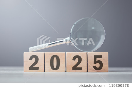Wooden block displaying '2025' placed beside a magnifying glass on a table, set against a gray background. Symbolizes a closer look at future plans, analysis, and preparation for the year ahead 119800378