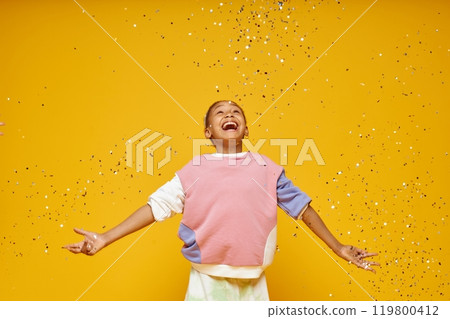 Medium shot of little excited Black girl throwing glittering confetti in air while standing against vivid orange background in studio, copy space 119800412