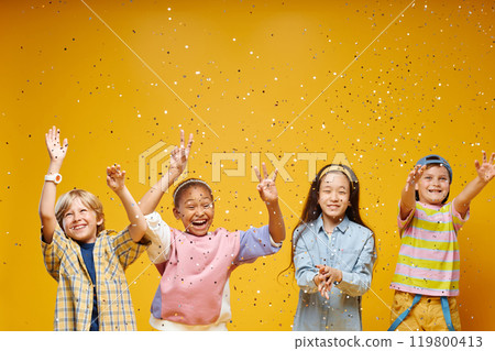 Medium shot of diverse group of four little excited kids jumping in air to catch confetti while standing in line against vivid orange background in studio, copy space 119800413