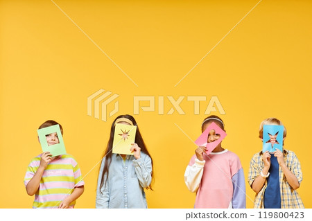 Medium full shot of four little kids covering faces with colorful paper crafts, while looking at camera through geometric shapes cut out on paper sheets on yellow background in studio 119800423
