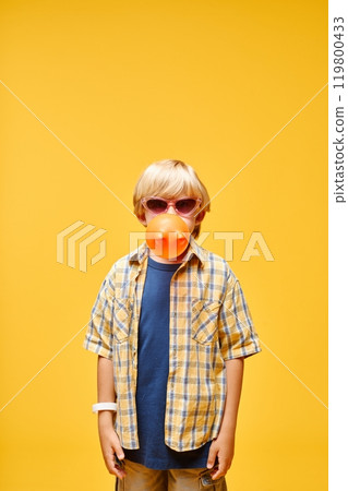 Vertical shot of little boy in sunglasses blowing bubblegum bubble while standing against yellow background in studio, copy space 119800433