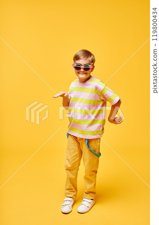 Vertical shot of smiling preteen boy fooling around while dancing in three pairs of colorful sunglasses against yellow background in studio, copy space 119800434