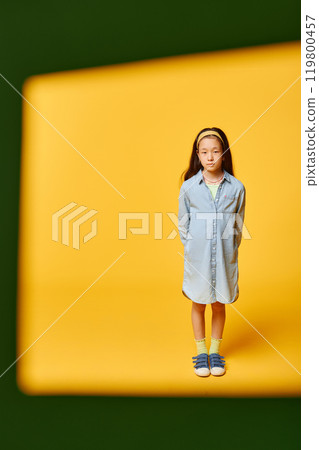 Vertical shot through green frame of cute preteen girl wearing denim shirt dress while standing against yellow background in studio, copy space Vertical shot through green frame of cute preteen girl wearing denim shirt dress while standing against yellow background in studio, copy space 119800457