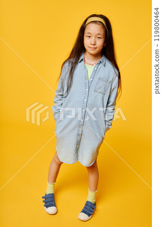 Vertical shot adorable little Asian girl wearing denim shirt dress keeping hands in pockets while standing against yellow background smiling at camera in studio, copy space Vertical shot adorable little Asian girl wearing denim shirt dress keeping hands in pockets while standing against yellow background smiling at camera in studio, copy space 119800464