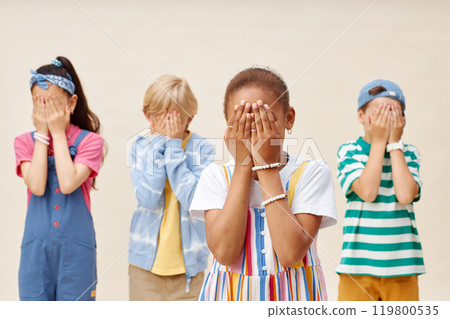 Medium shot of cute little Black girl in colorful clothes covering face with both hands as if counting, while playing hide and seek game with friends against neutral background in studio Medium shot of cute little Black girl in colorful clothes covering face with both hands as if counting, while playing hide and seek game with friends against neutral background in studio 119800535