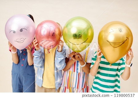 Medium shot of four little kids wearing colorful clothes holding balloons with funny drawings while covering faces and hiding from camera on neutral background in studio, copy space 119800537