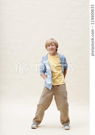 Vertical shot of cheerful little boy wearing blue zip up hoodie keeping hands in pockets while showcasing casual outfit for active games on neutral background in studio, copy space 119800633