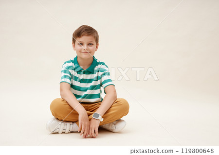 Cute little boy wearing vibrant striped polo t shirt sitting cross legged while smiling at camera on neutral background in studio, copy space 119800648