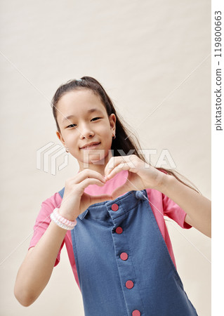 Vertical portrait shot of cute Asian girl showing heart gesture smiling at camera expressing love and appreciation while posing against neutral studio background, copy space Vertical portrait shot of cute Asian girl showing heart gesture smiling at camera expressing love and appreciation while posing against neutral studio background, copy space 119800663