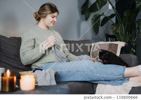 Pregnant woman knitting with black cat beside, preparing handmade baby items. Cozy nesting, relaxation, and mindful pregnancy. 119800666
