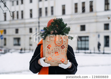A person carrying a festive gift bag filled with evergreen branches in a snowy park, highlighting eco-friendly and minimalist holiday trends. 119800669