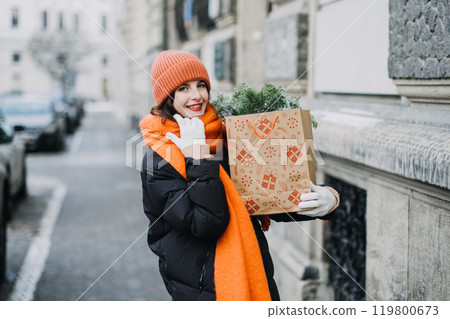 Christmas routine. Woman holding a festive shopping bag filled with pine branches, smiling in winter attire during Christmas season shopping Christmas routine. Woman holding a festive shopping bag filled with pine branches, smiling in winter attire during Christmas season shopping 119800673