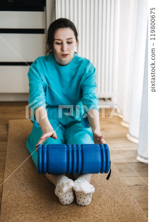 Young woman in blue clothing using foam roller for myofascial release and muscle tension relief. Physical therapy, relaxation, and self-care concept 119800675