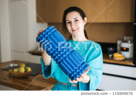Woman in casual blue outfit showing a foam roller in home, highlighting home fitness, self-care, and muscle recovery routines 119800676
