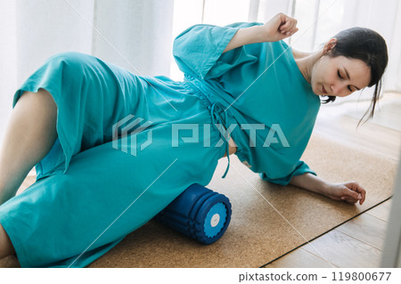Young woman in blue clothing using foam roller for myofascial release and muscle tension relief. Physical therapy, relaxation, and self-care concept 119800677