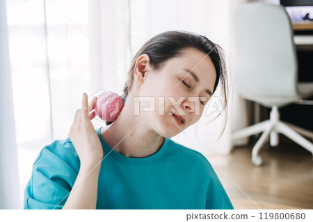 Woman practicing self-massage on her neck with a pink massage ball, focusing on stress relief and relaxation in a home setting 119800680