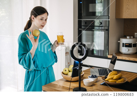 Young woman creating social media content on healthy lifestyle with fruits and juice in kitchen. Wellness influencer and food vlogging concept Young woman creating social media content on healthy lifestyle with fruits and juice in kitchen. Wellness influencer and food vlogging concept 119800683