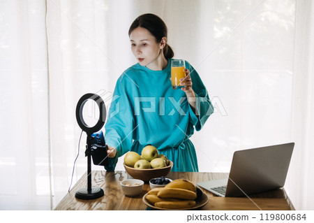 Young woman creating social media content on healthy lifestyle with fruits and juice in kitchen. Wellness influencer and food vlogging concept 119800684
