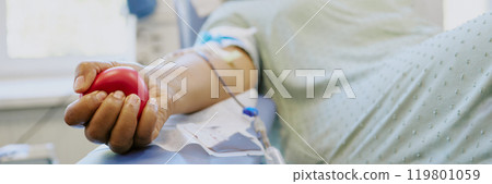 Close-up of a person donating blood at a medical clinic, holding a red squeeze ball. Intravenous equipment and hospital gown visible 119801059