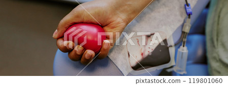 Hand holding heart-shaped squeeze ball while donating blood via intravenous drip. Close-up showcasing medical procedure and device used for blood transfusion 119801060