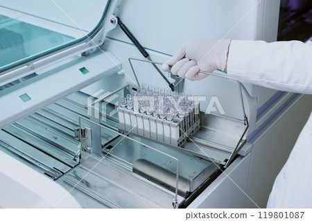 Close-up of gloved hand holding metal rack filled with multiple blood samples inside advanced laboratory equipment for testing and diagnostics 119801087