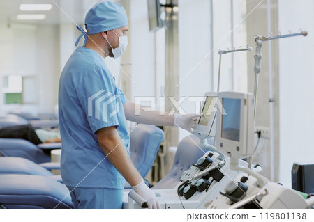 Technician wearing mask and scrubs adjusting medical equipment in hospital ward. Devices in focus with patient beds in background Technician wearing mask and scrubs adjusting medical equipment in hospital ward. Devices in focus with patient beds in background 119801138