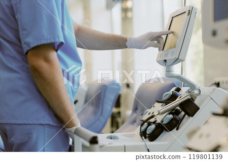 Medical professional in scrubs adjusting touchscreen on medical device in clinical setting while interacting with equipment. Focus on hand and device while working Medical professional in scrubs adjusting touchscreen on medical device in clinical setting while interacting with equipment. Focus on hand and device while working 119801139